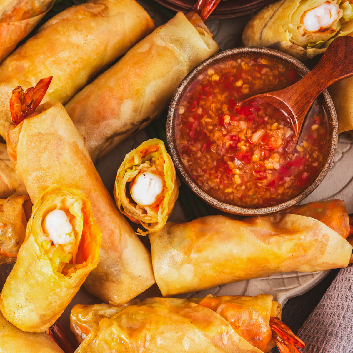 A party platter of golden fried shrimp spring rolls with tails sticking out the end of the wrappers next to a bowl of bright Thai chili sauce.