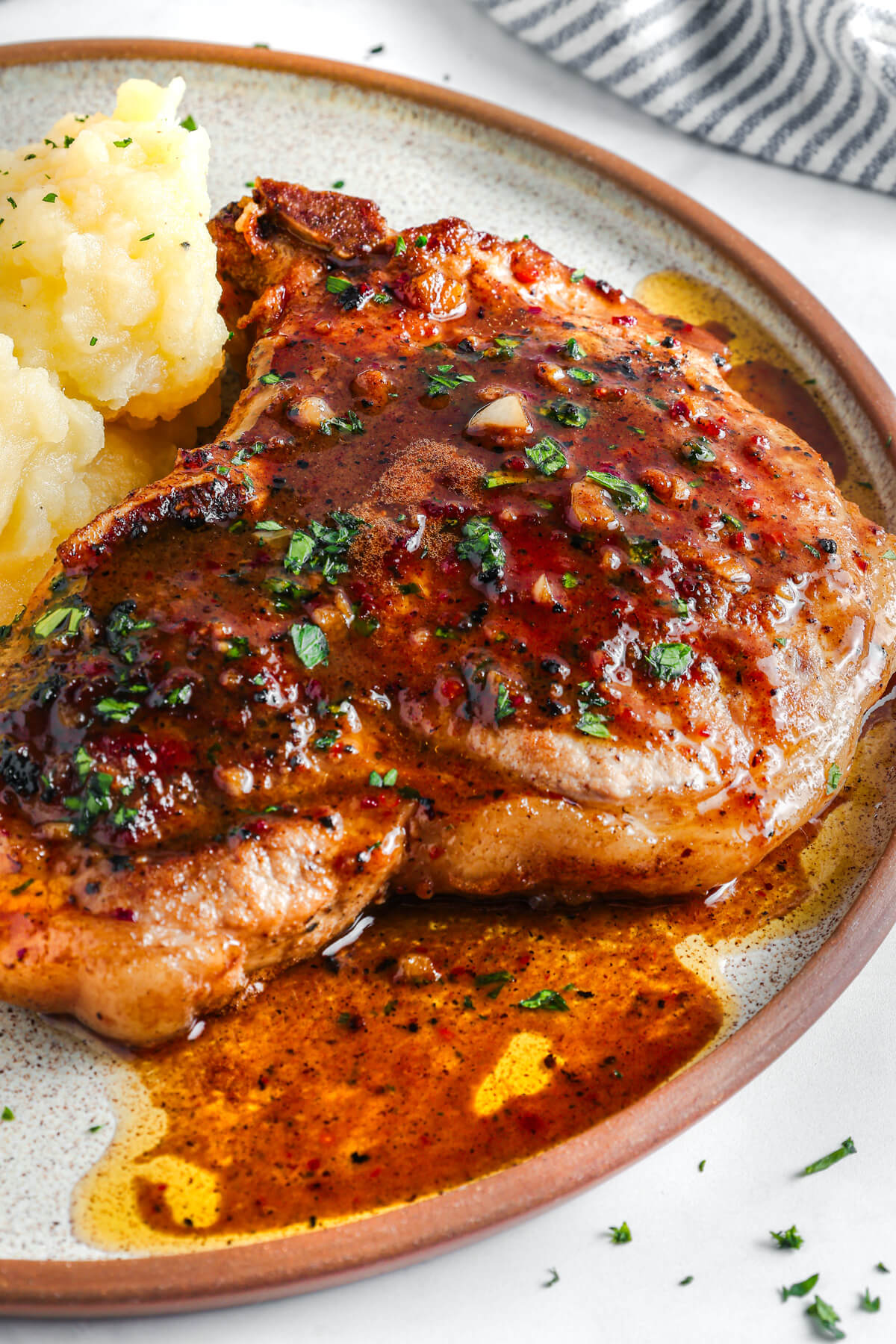 A perfectly seared brown sugar pork chop covered in luxurious brown sauce, parsley, and garlic on a dinner plate with mashed potatoes.