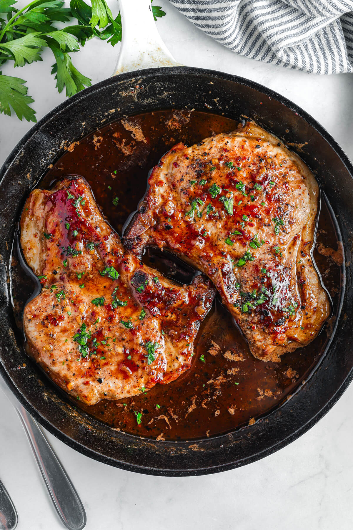 Two perfectly seared brown sugar pork chops covered in luxurious brown sauce, parsley, and garlic in a cast iron skillet.