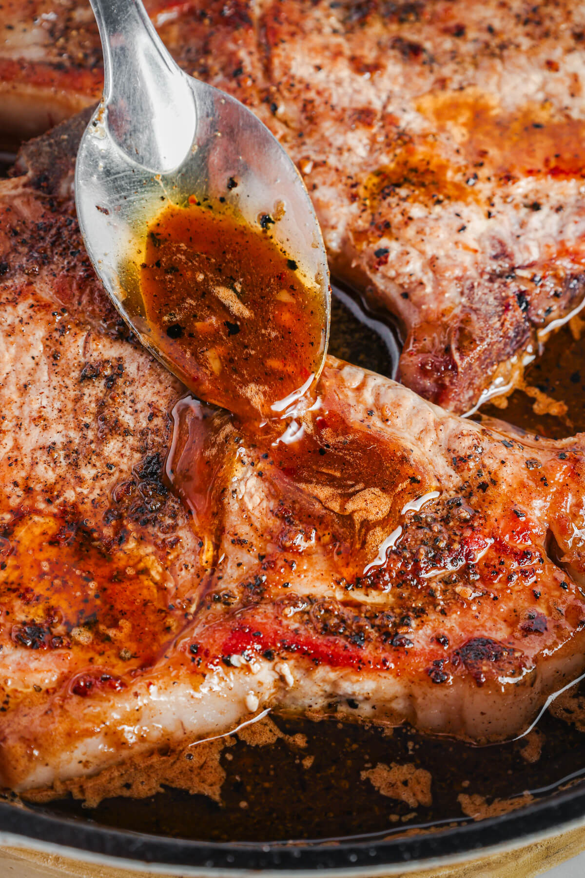 Two perfectly seared brown sugar pork chops being covered in luxurious brown sauce, parsley, and garlic in a cast iron skillet.