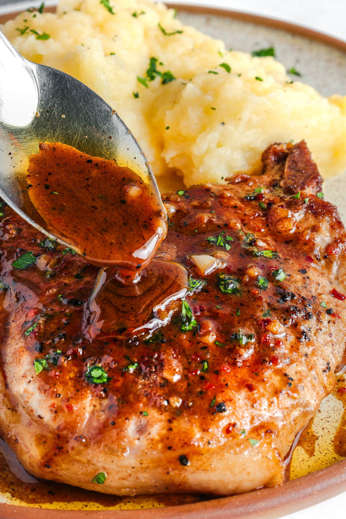 A perfectly seared brown sugar pork chop being covered in luxurious brown sauce, parsley, and garlic on a dinner plate with mashed potatoes.