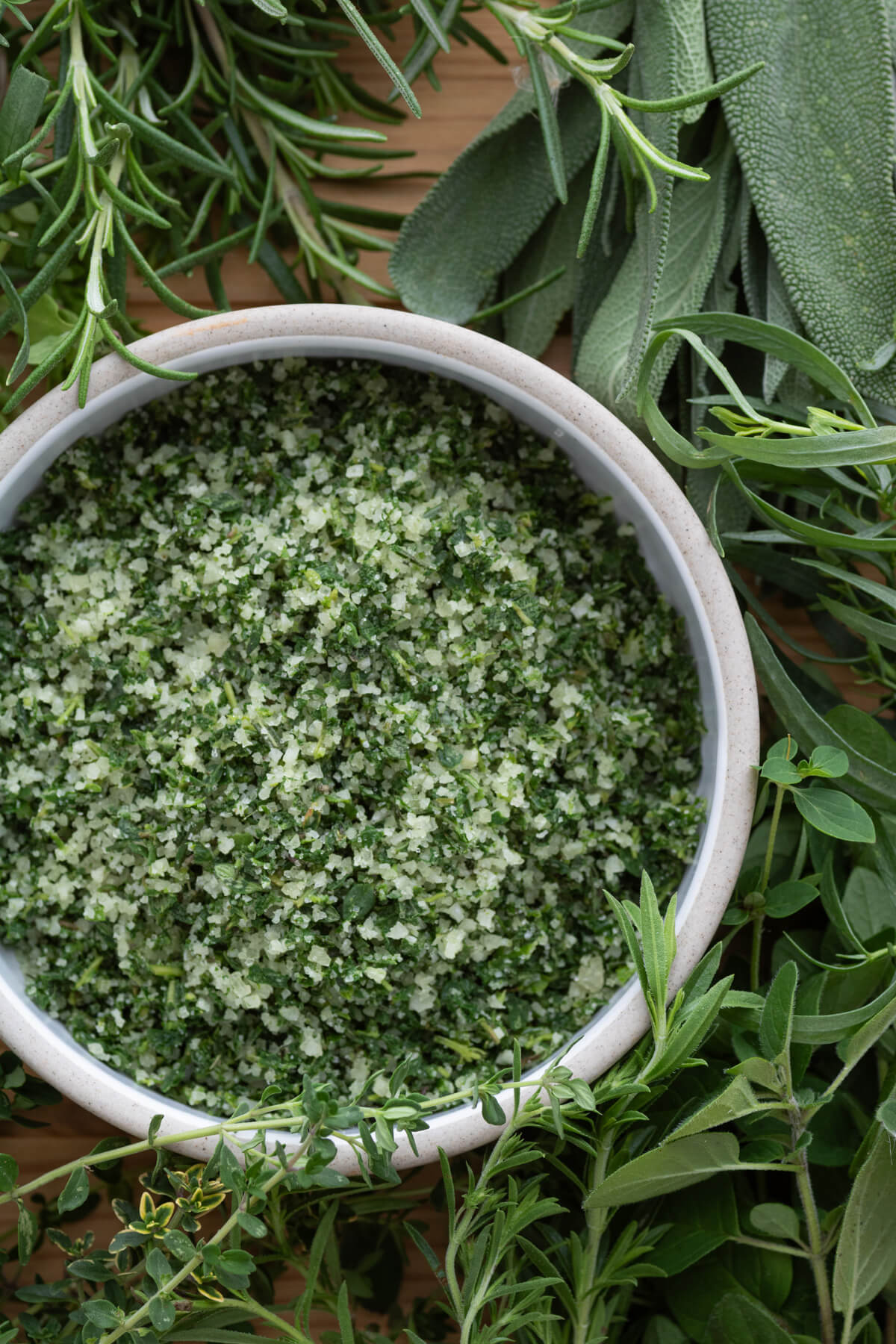 A round dish containing vibrant green herb salt surrounded by green herbs.