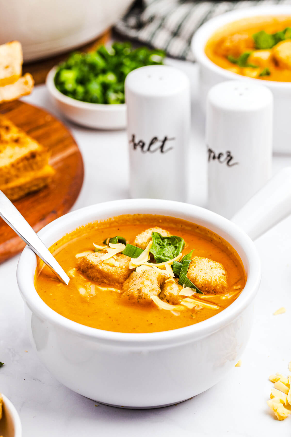 A bowl of bright orange, smooth and creamy Roasted Red Pepper Soup garnished with fresh basil, grated cheese and croutons with a spoon.