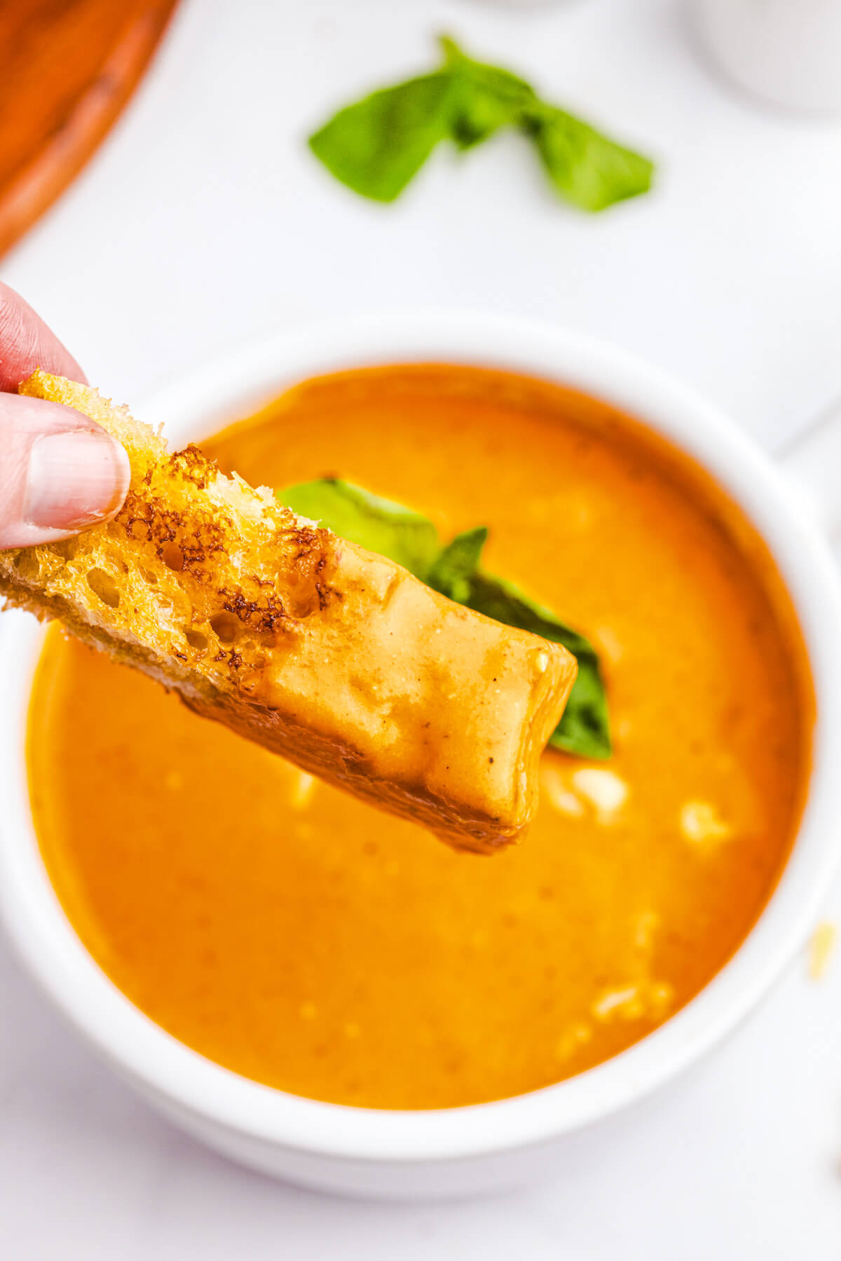 A piece of crusty bread coated in roasted red pepper soup above a bowl of the same soup.
