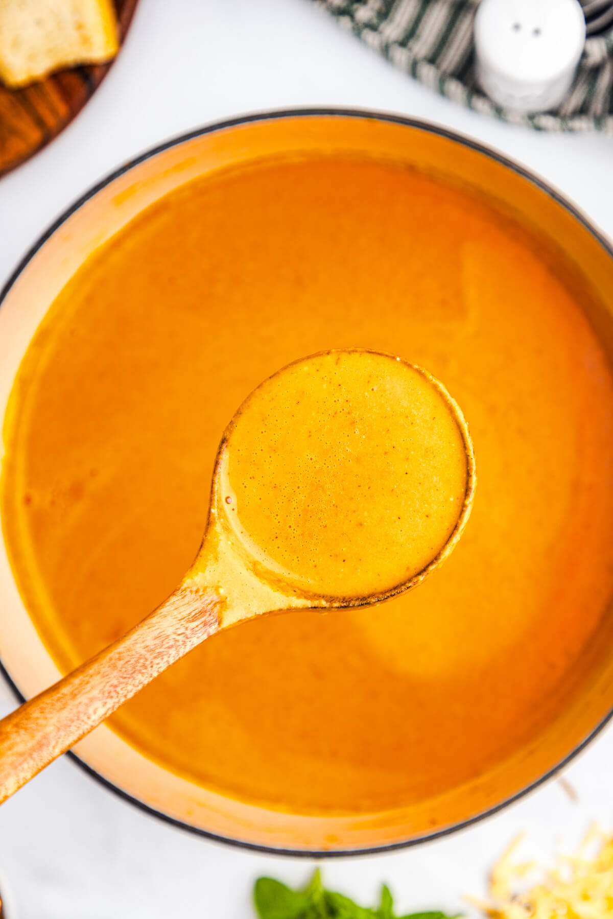 A wooden spoon full of roasted red pepper soup above a pot filled with the same vibrant orange soup.