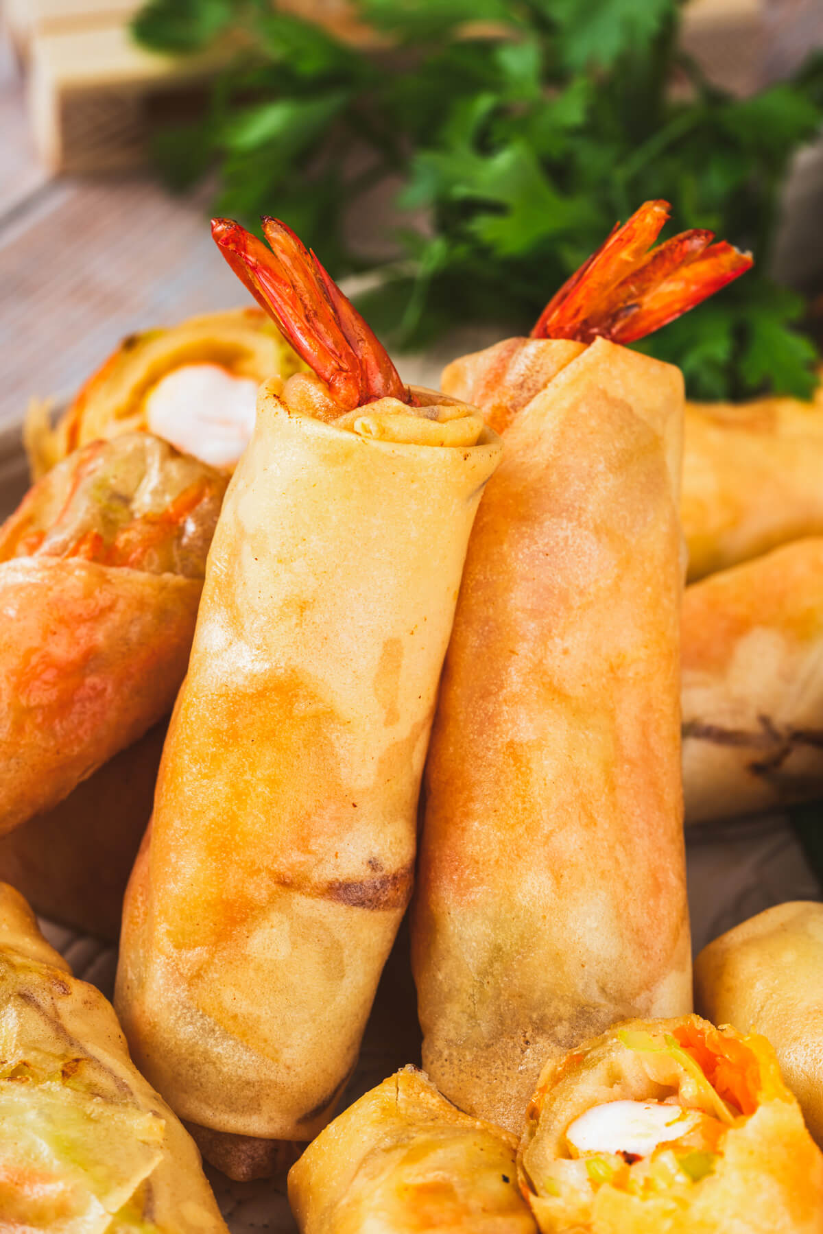 Two golden fried shrimp spring rolls in wrappers with tails sticking out lean against a pile of more spring rolls.