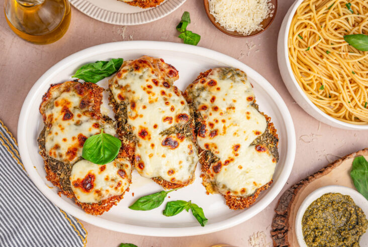 Three Baked Pesto Chicken Parmesans covered in green pesto, red tomato sauce, and gooey melted mozzarella cheese on a white platter garnished with fresh basil.