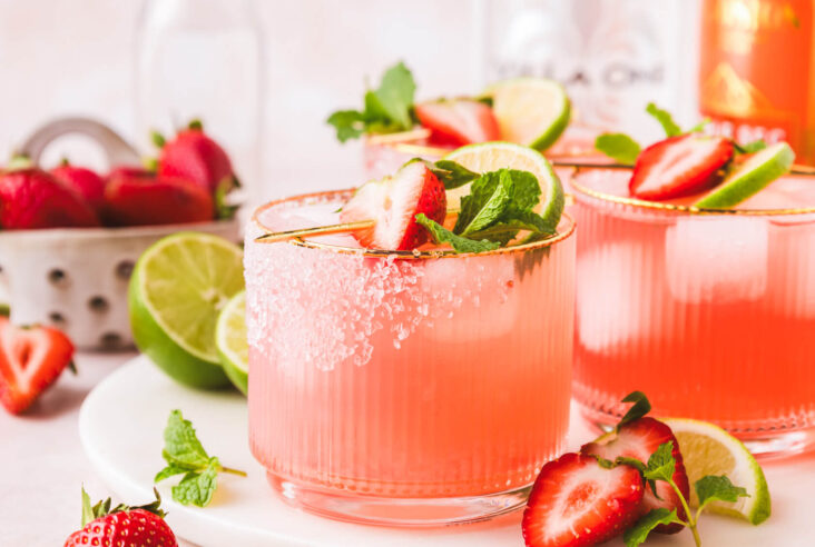 Two rocks cocktail glasses with a sugar rims containing vibrant pink strawberry margarita on ice garnished with a half strawberry, mint leaves, and a lime wheel.