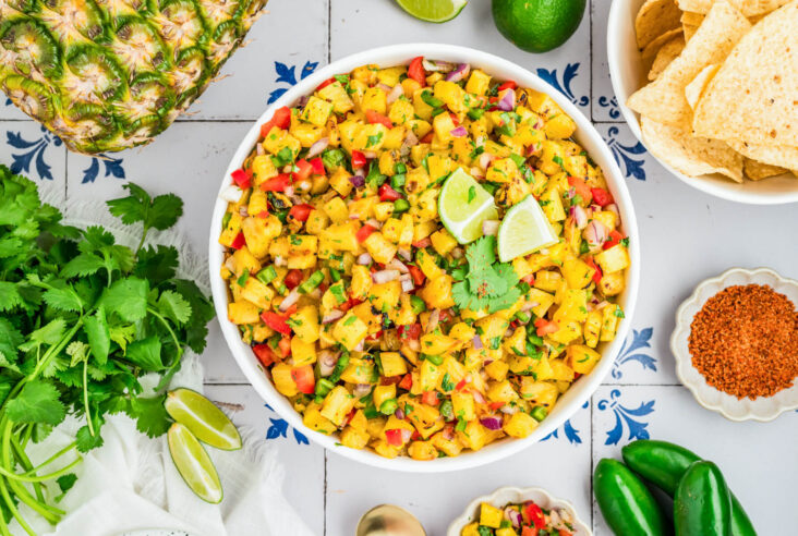 A white a bowl containing vibrant Grilled Pineapple Salsa with red bell pepper, jalapeno pepper, green cilantro, grilled pineapple, red onion, and lime wedges.