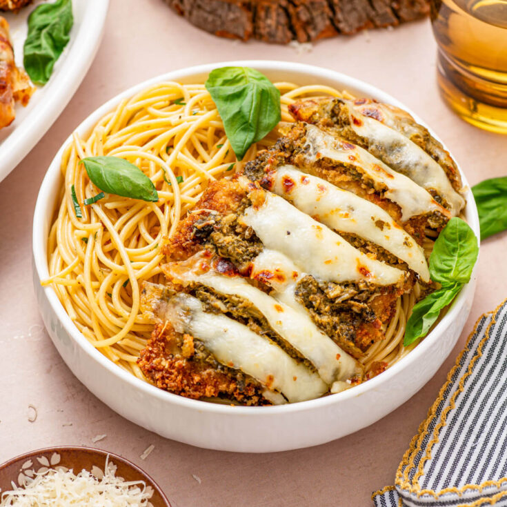 Baked and sliced Pesto Chicken Parmesan covered in green pesto, red tomato sauce, and gooey melted mozzarella cheese in a bowl of spaghetti.