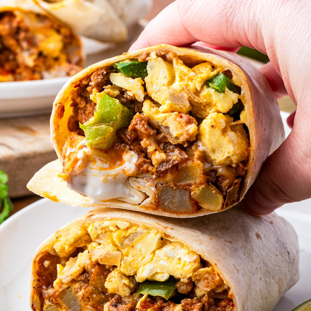 A hand holds half of a sliced Mexican Breakfast Burrito with spicy red chorizo, soft scrambled eggs, melted cheese and vegetables.