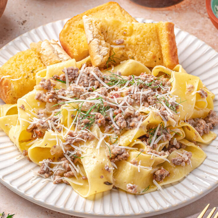 A white pasta bowl containing thick pappardelle in a creamy white Ragù Bianco with a side of garlic bread.