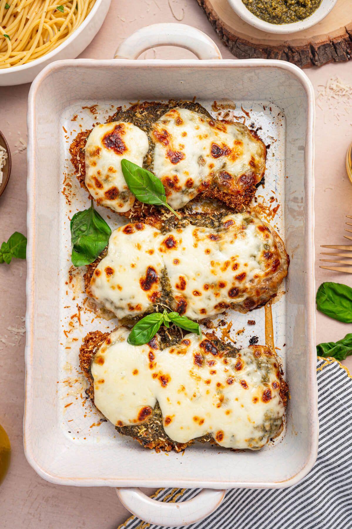 Three Baked Pesto Chicken Parmesans covered in green pesto, red tomato sauce, and gooey melted mozzarella cheese in a white baking dish garnished with fresh basil.