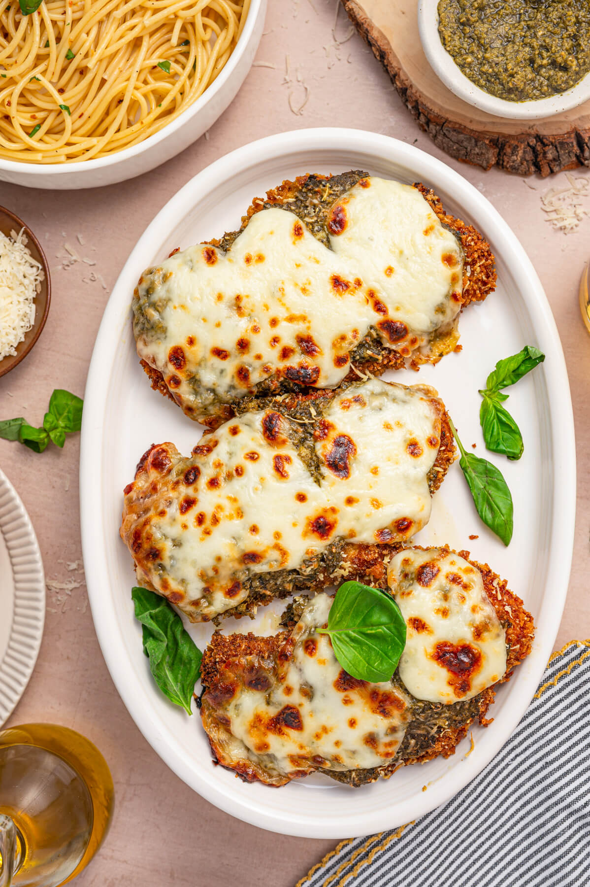 Three Baked Pesto Chicken Parmesans covered in green pesto, red tomato sauce, and gooey melted mozzarella cheese on a white platter garnished with fresh basil.