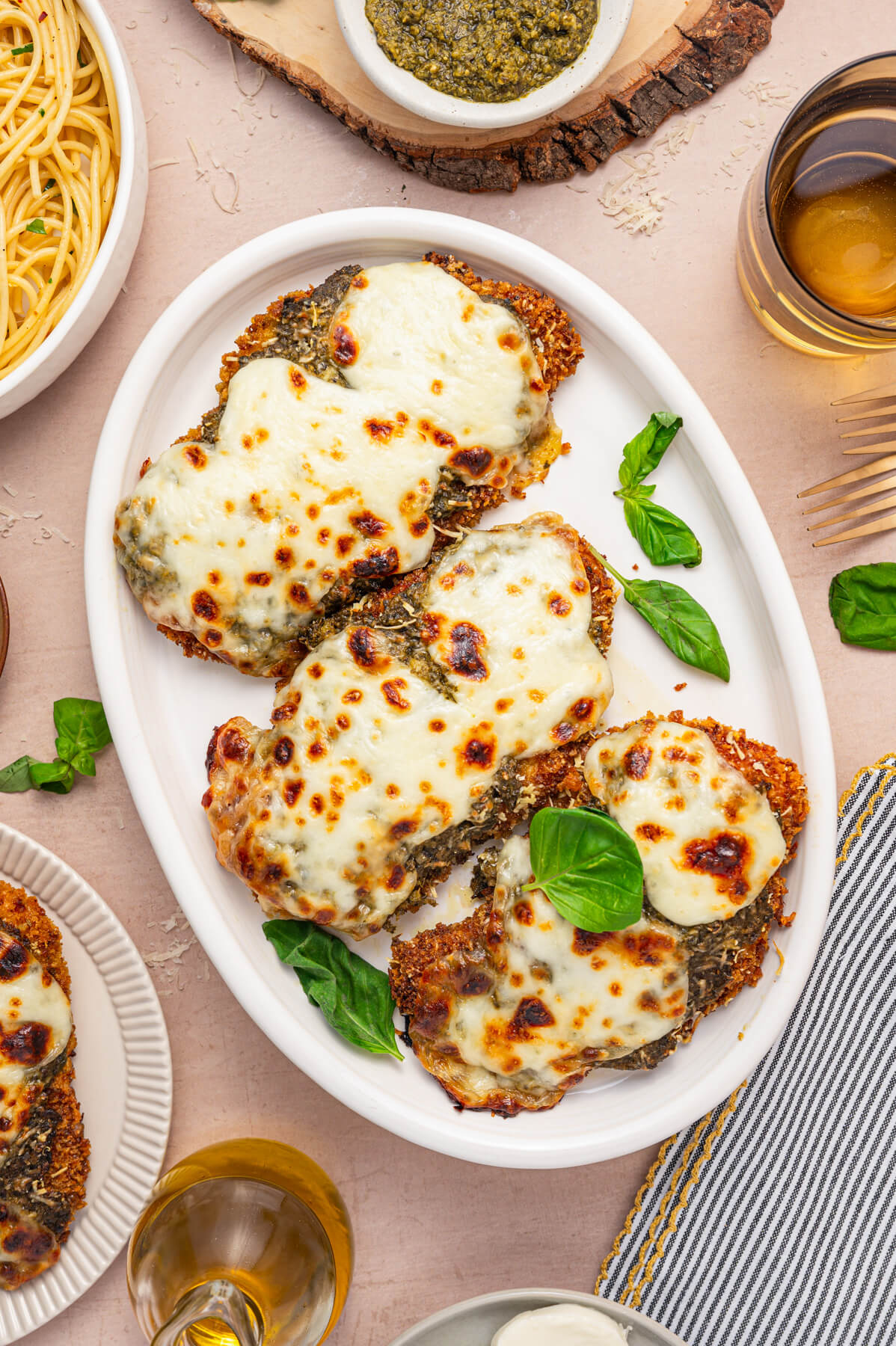 Three Baked Pesto Chicken Parmesans covered in green pesto, red tomato sauce, and gooey melted mozzarella cheese on a white platter garnished with fresh basil.