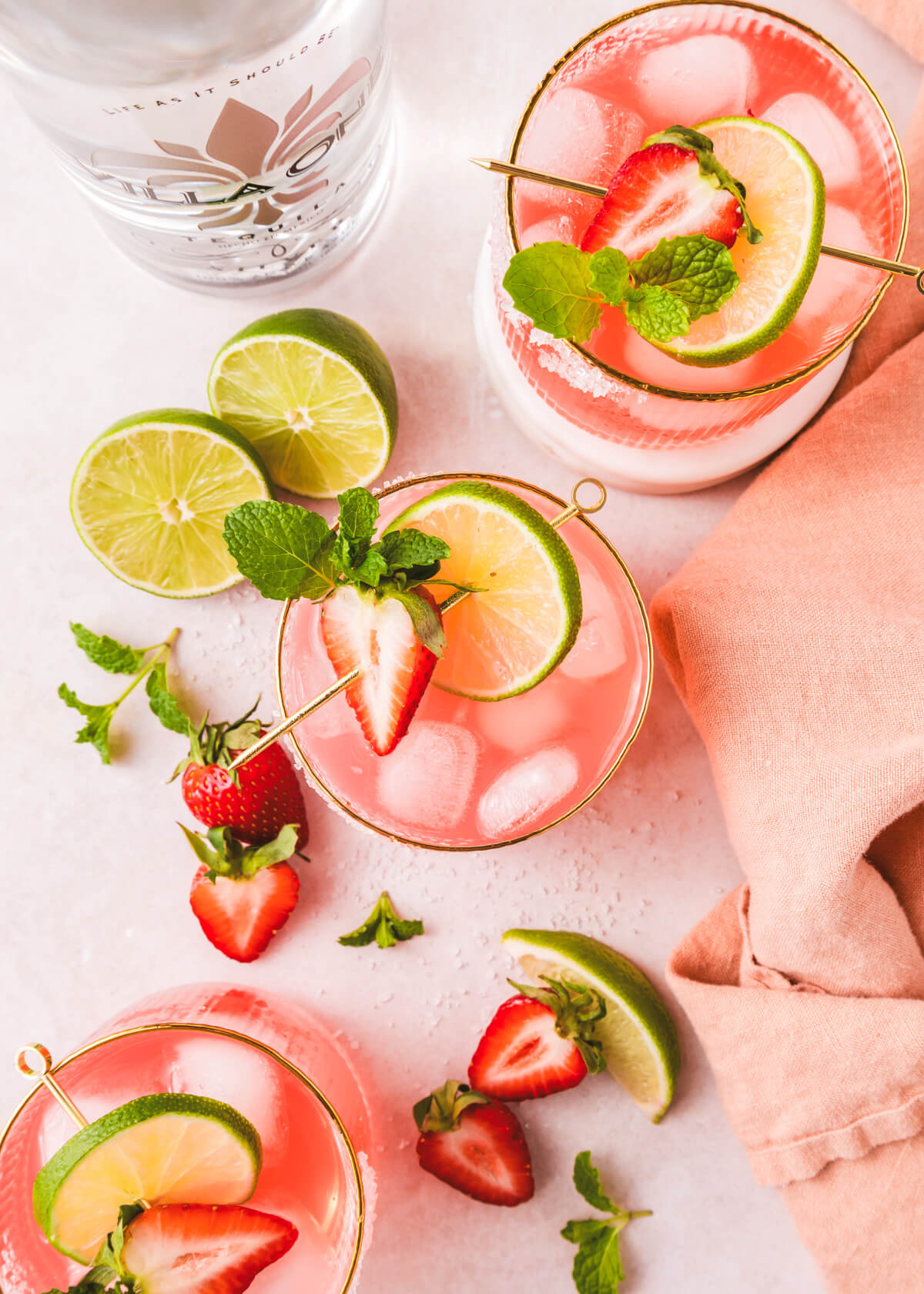 Three rocks cocktail glasses with sugar rims containing vibrant pink strawberry margarita on ice garnished with a half strawberry, mint leaves, and lime wheels.