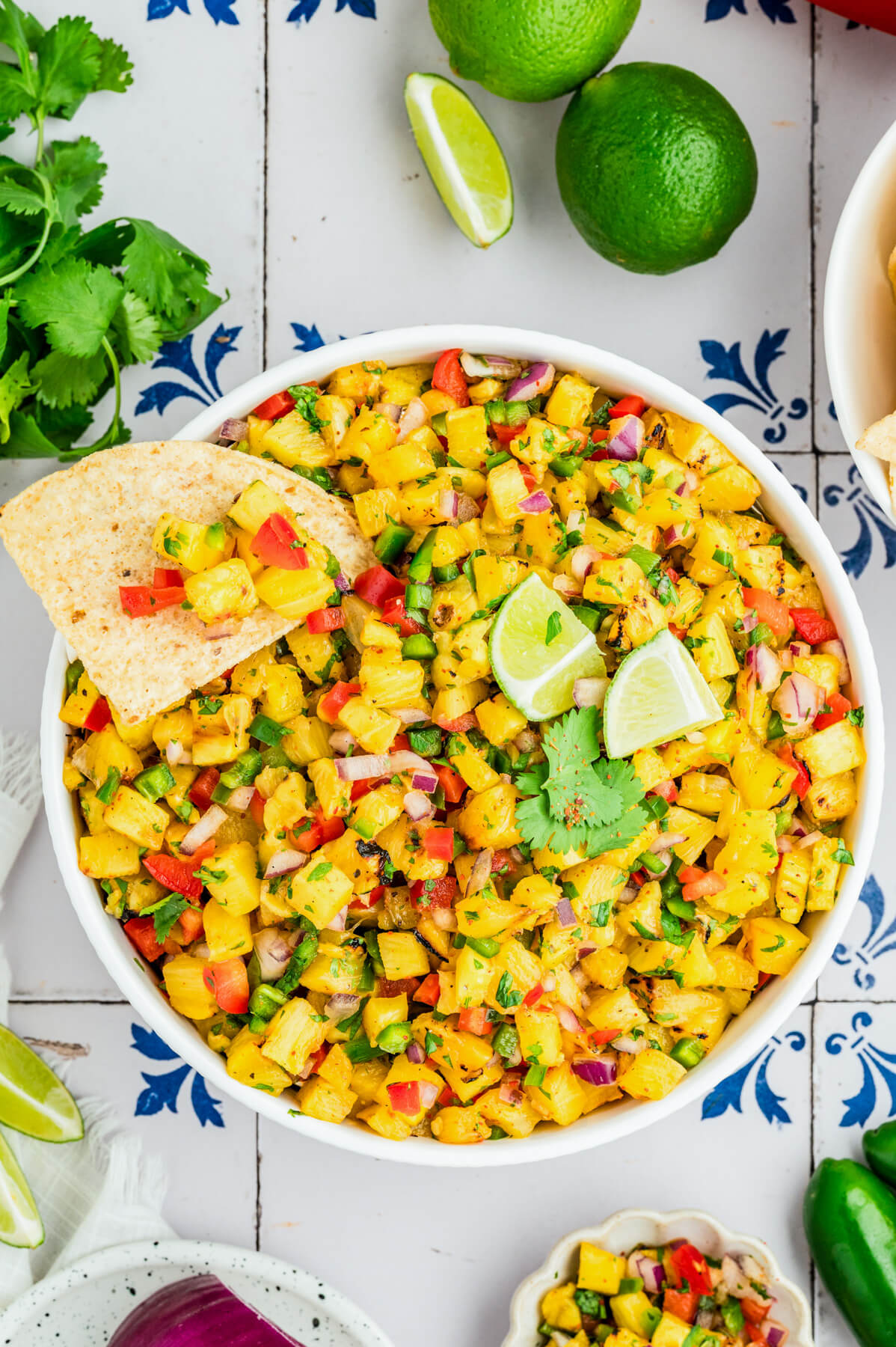 A white a bowl containing vibrant Grilled Pineapple Salsa with red bell pepper, jalapeno pepper, green cilantro, grilled pineapple, red onion, and lime wedges.