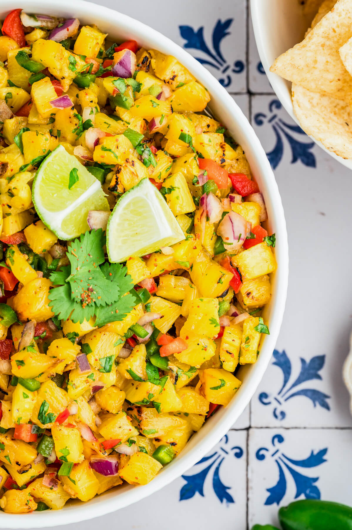 A white a bowl containing vibrant Grilled Pineapple Salsa with red bell pepper, jalapeno pepper, green cilantro, grilled pineapple, red onion, and lime wedges.