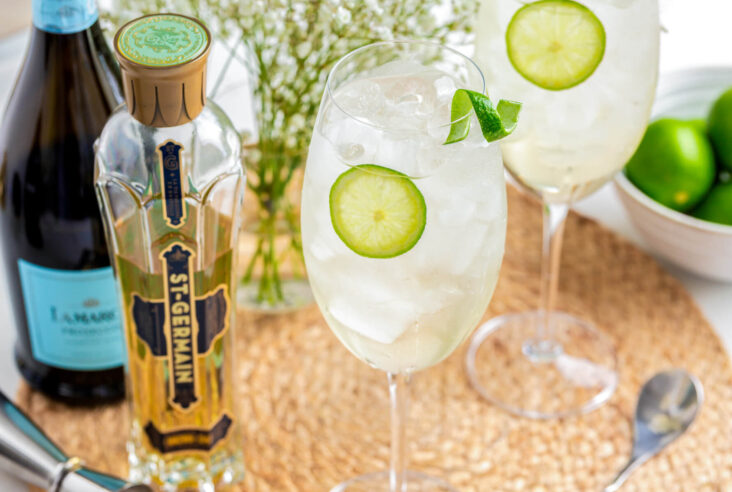 Two icy St. Germain Spritz cocktails in large wine glasses garnished with slices of lime.