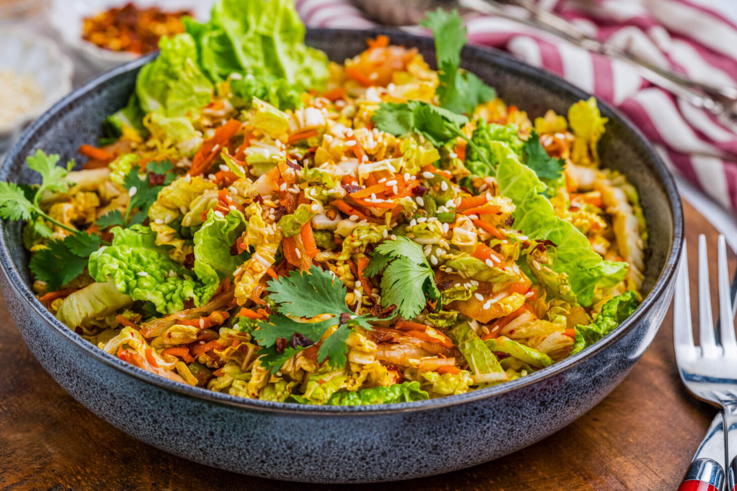A bowl of vibrant kimchi slaw with green cilantro, sesame seeds, and shredded carrots on a wooden board.