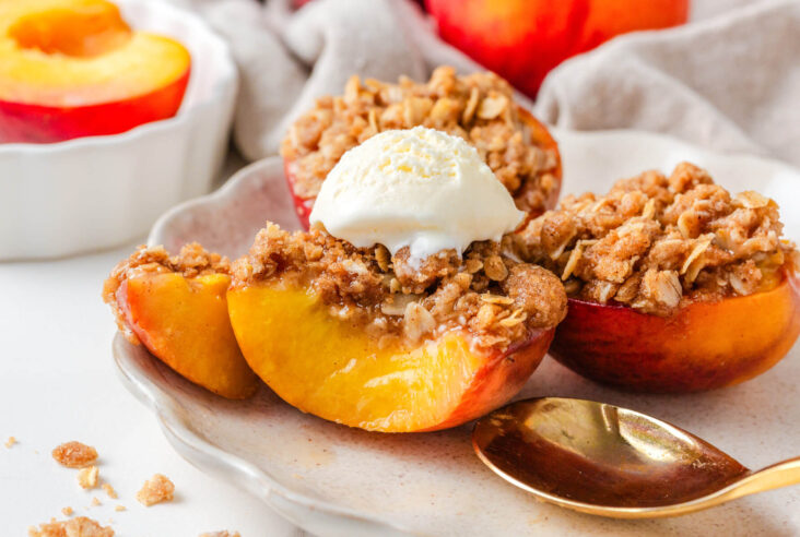 Four baked peaches topped with buttery oat crumble and vanilla ice cream on a white plate.