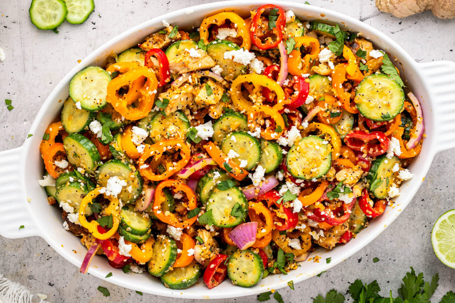 A white oval serving dish containing a vibrant Cucumber Sweet Pepper Salad with chicken, sesame seeds, feta cheese, red onions, cilantro in a honey lime dressing.