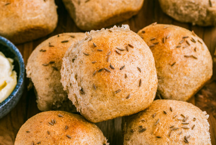 A group of golden baked rye dinner rolls dotted with caraway seeds on a wooden board.