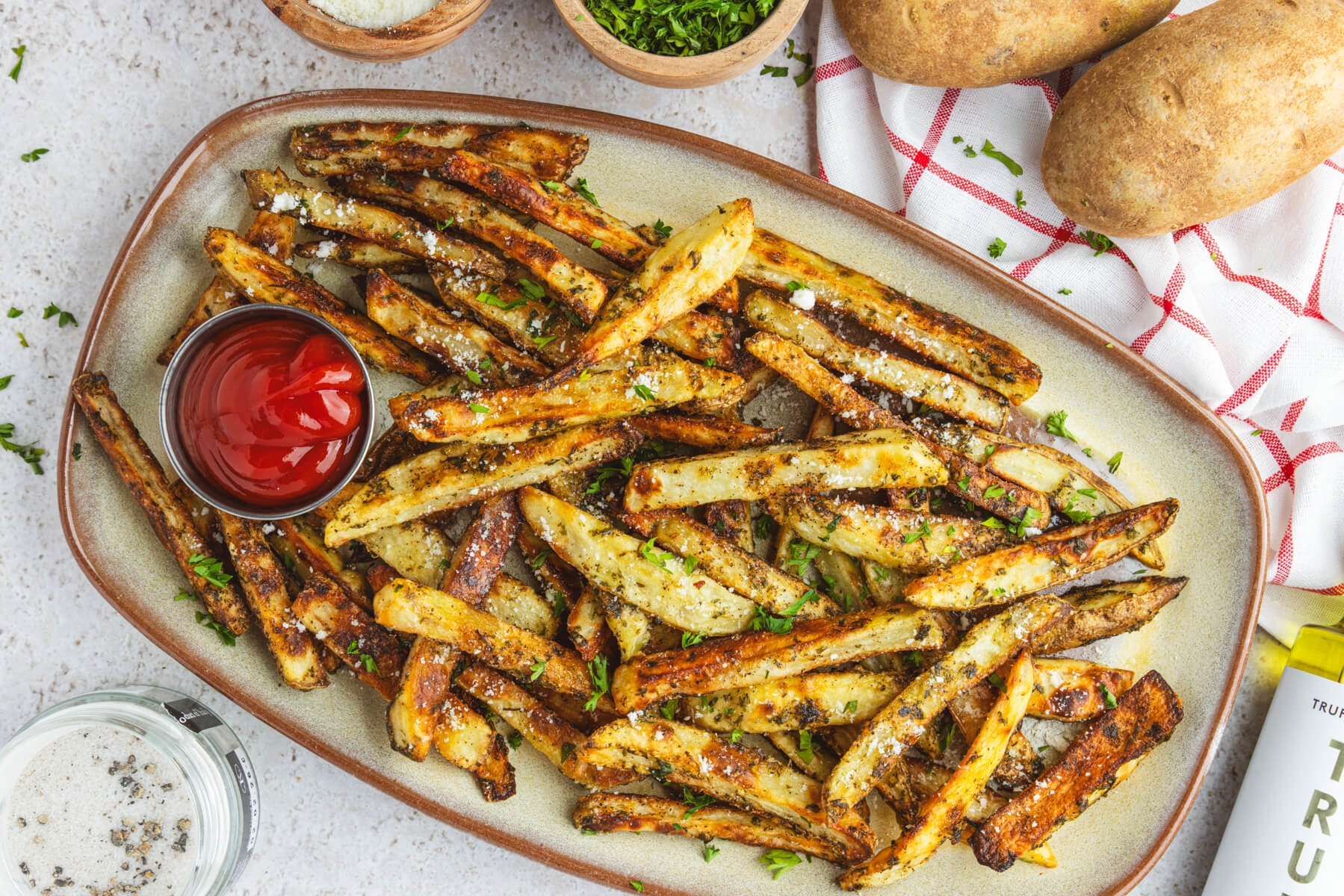 A plate of golden brown oven baked truffle fries dotted with parsley and black truffle salt with a container of ketchup.