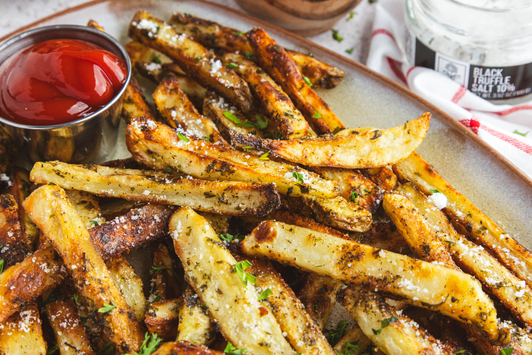 A plate of golden brown oven baked truffle fries dotted with parsley and black truffle salt with a container of ketchup.