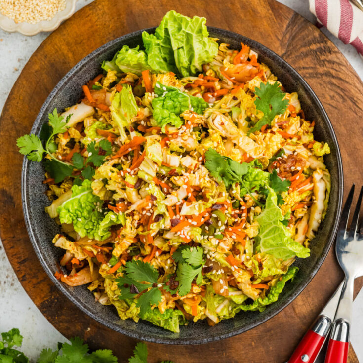 A bowl of vibrant kimchi slaw with green cilantro, sesame seeds, and shredded carrots on a wooden board.