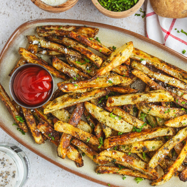A plate of golden brown oven baked truffle fries dotted with parsley and black truffle salt with a container of ketchup.