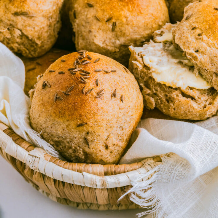 A basket filled with golden baked rye dinner rolls dotted with caraway seeds.