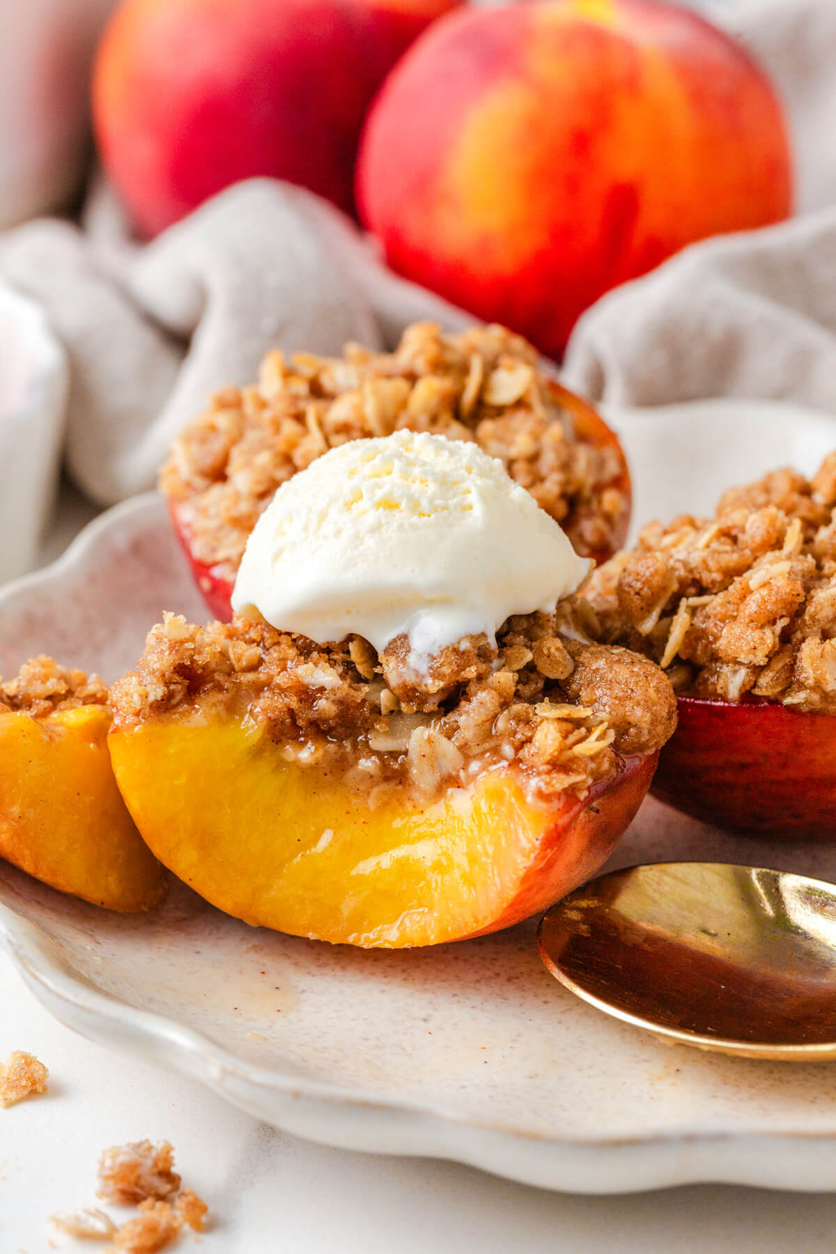 Four baked peaches topped with buttery oat crumble and vanilla ice cream on a white plate.