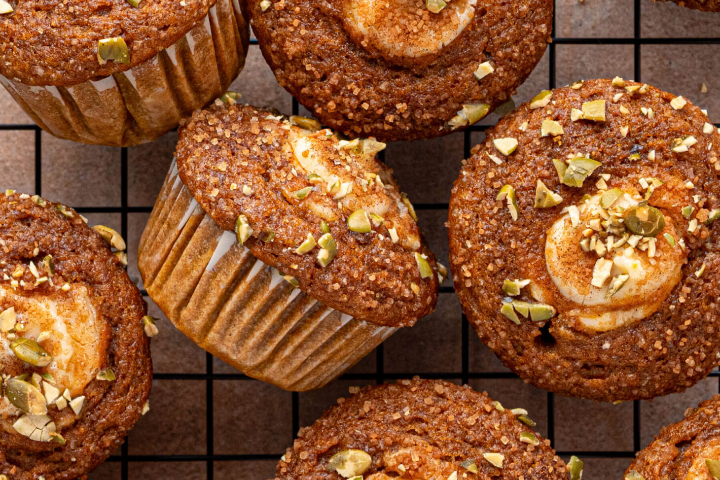 A group of golden baked Pumpkin Cream Cheese Muffins with a cream cheese, sugar, and pumpkin seed topping on a baking rack.