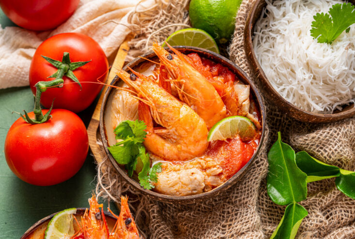 A half coconut bowl filled with vibrant Tom Yum soup, succulent shrimp, fresh herbs, noodles, and a wedge of lime surrounded by recipe ingredients.