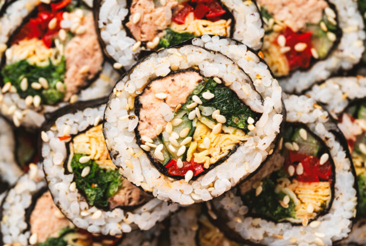 A close up photo of a plate of Tuna Kimbap roll featuring canned tuna in mayonnaise, egg omelet, spinach, and red bell pepper wrapped in seaweed and rice.