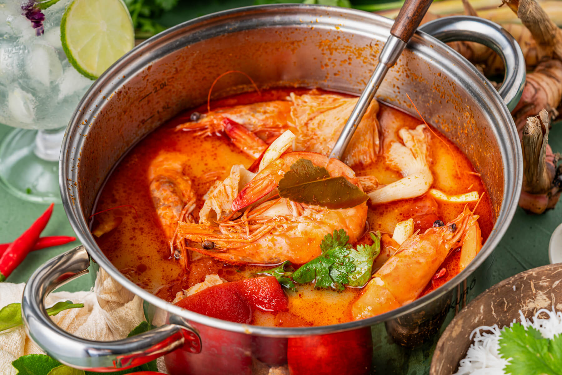 A soup pot filled with vibrant Tom Yum soup, succulent shrimp, mushrooms, tomatoes, noodles, and fresh herbs.