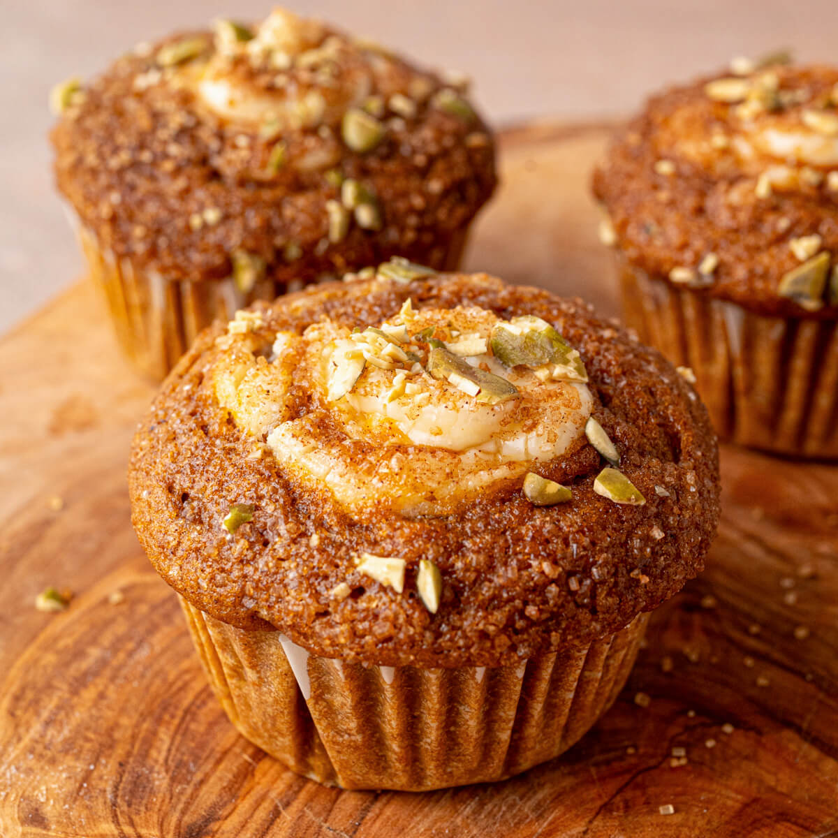 A group of golden baked Pumpkin Cream Cheese Muffins with a cream cheese, sugar, and pumpkin seed topping on a wooden board.