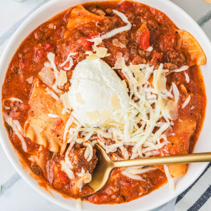 A spoon in a white soup bowl full of Slow Cooker Lasagna soup topped with ricotta, mozzarella, and Parmesan cheese.