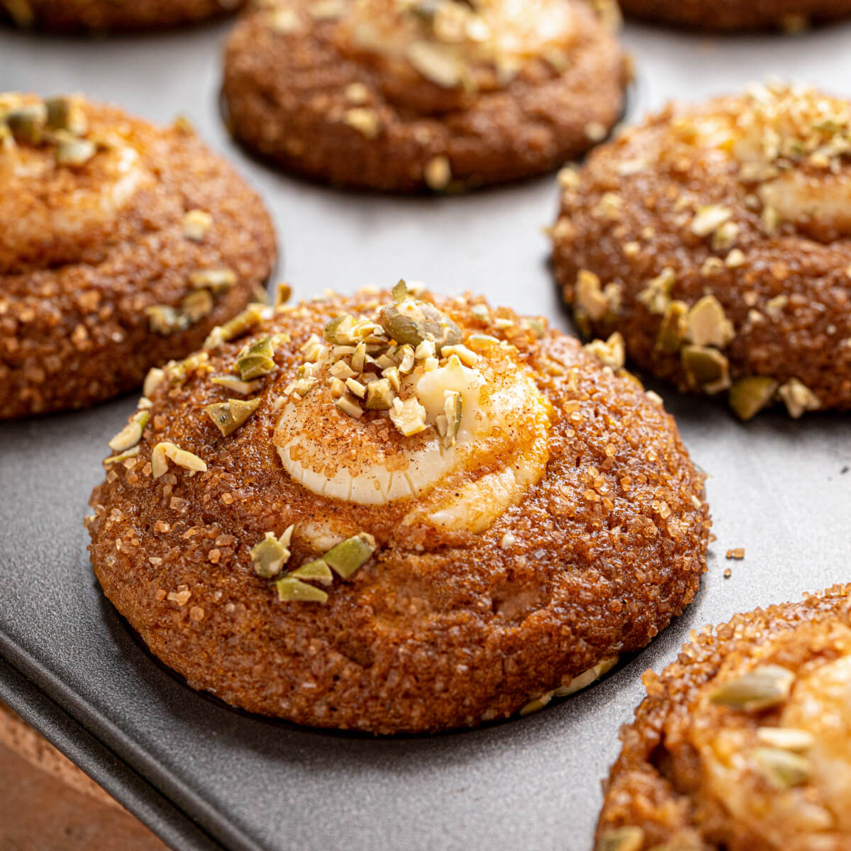 A group of golden baked Pumpkin Cream Cheese Muffins with a cream cheese, sugar, and pumpkin seed topping in a muffin tin.
