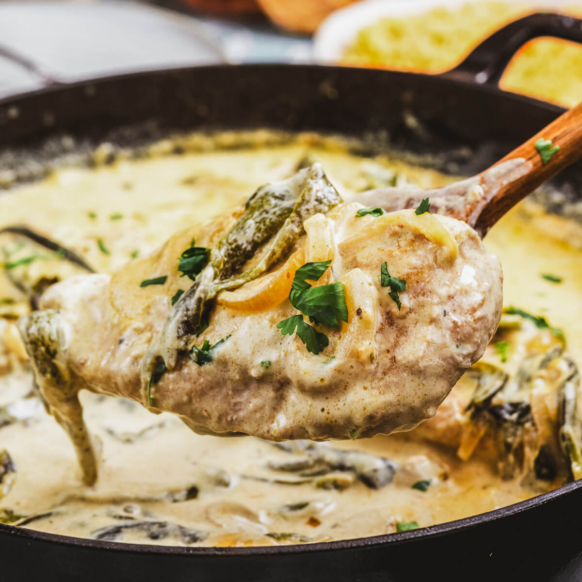 A wooden spoon holds a chicken breast covered in creamy rajas poblanas over a skillet of the same.