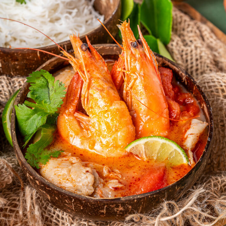 A half coconut bowl filled with vibrant Tom Yum soup, succulent shrimp, fresh herbs, noodles, and a wedge of lime surrounded by recipe ingredients.