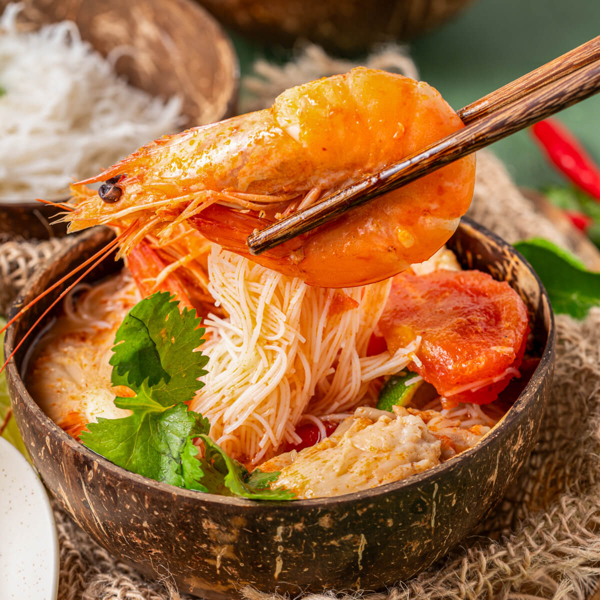 A pair of wooden chopsticks hold a large prawn over a bowl of vibrant red Tom Yum Soup.