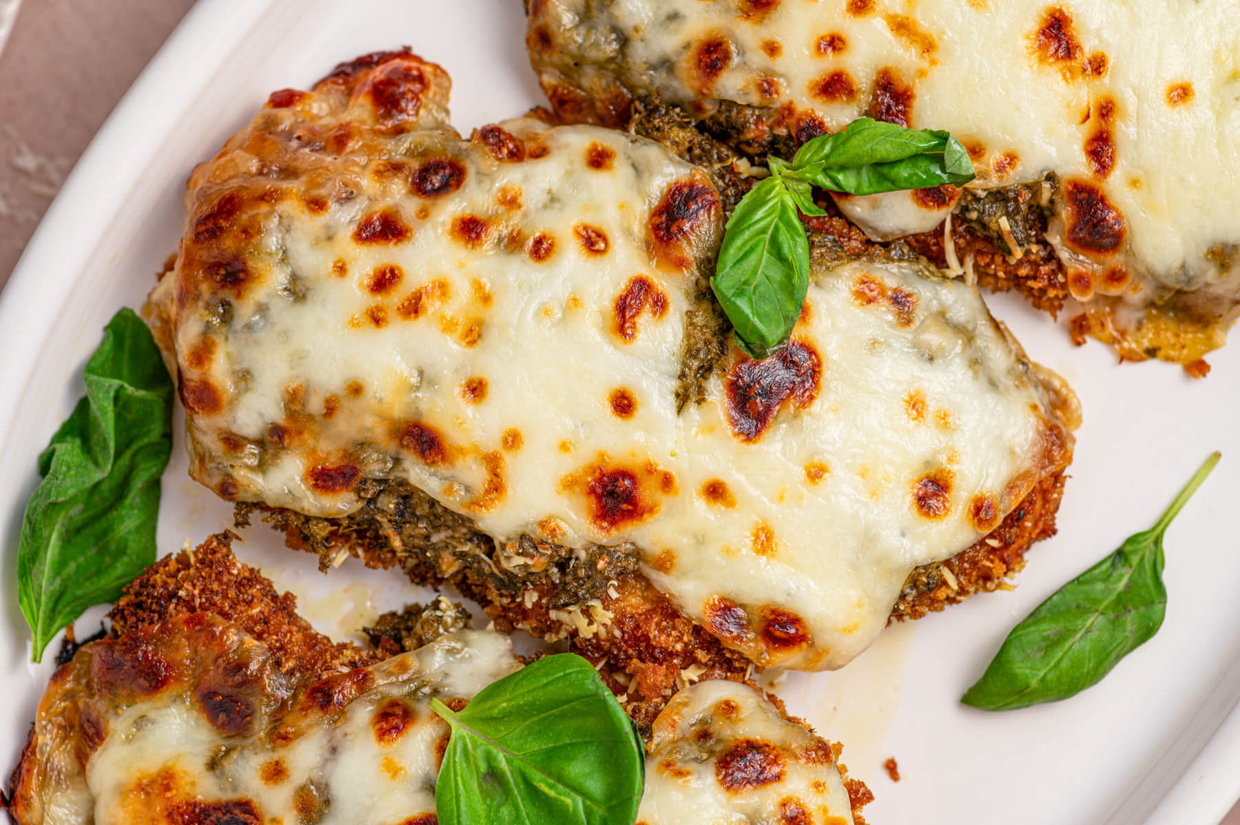 Three Baked Pesto Chicken Parmesans covered in green pesto, red tomato sauce, and gooey melted mozzarella cheese on a white platter garnished with fresh basil.