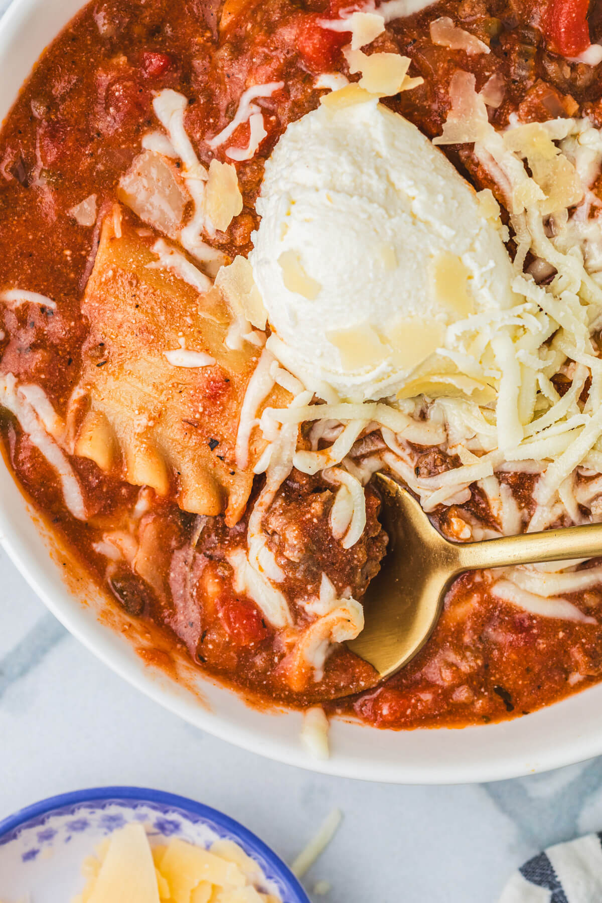 A spoon in a white soup bowl full of Slow Cooker Lasagna soup topped with ricotta, mozzarella, and Parmesan cheese.