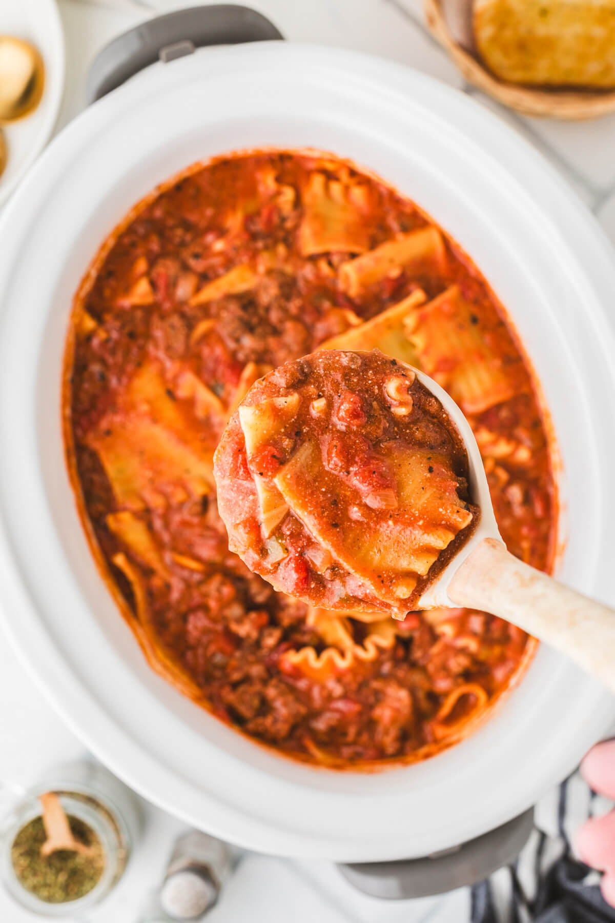 A full ladle of soup floats above a white oval slow cooker filled with meaty lasagna soup.