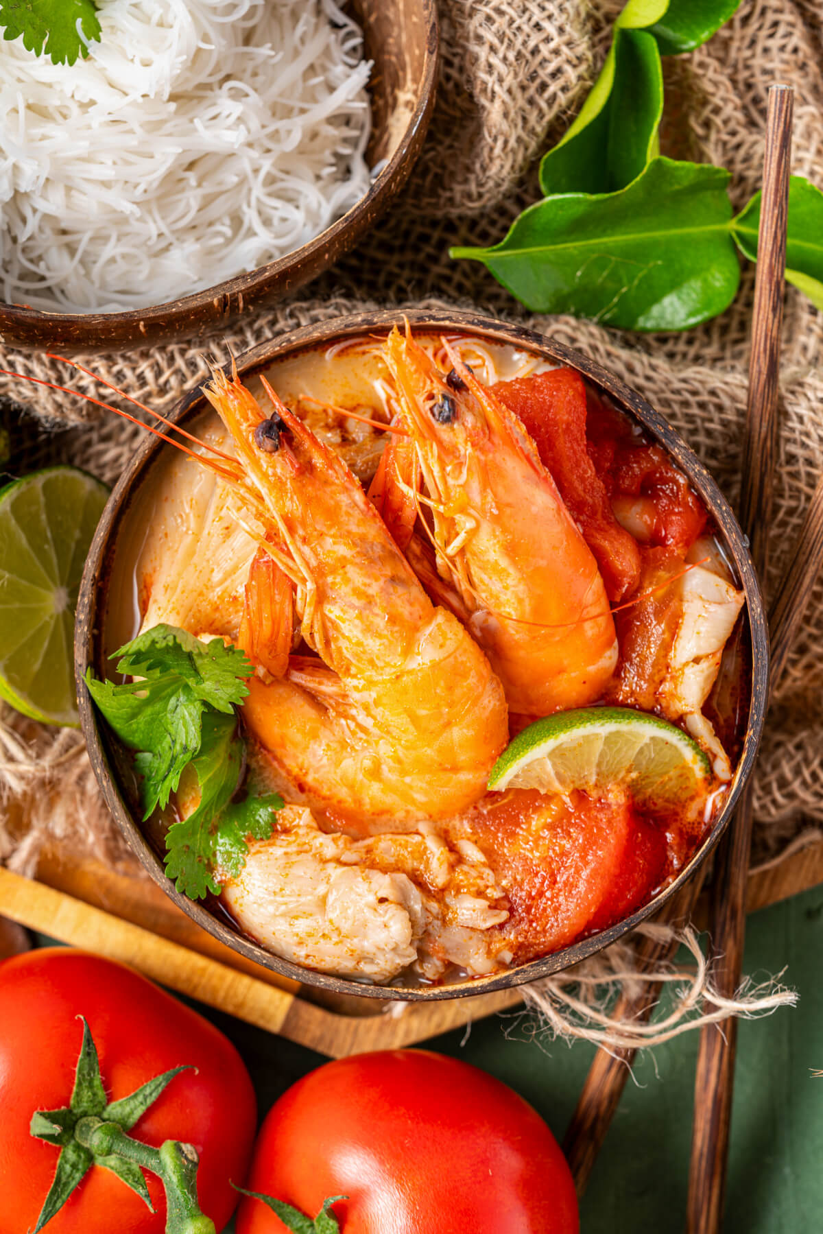 A half coconut bowl filled with vibrant Tom Yum soup, succulent shrimp, fresh herbs, noodles, and a wedge of lime.