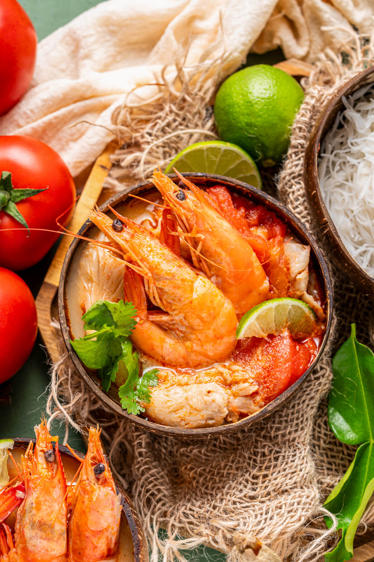 A half coconut bowl filled with vibrant Tom Yum soup, succulent shrimp, fresh herbs, noodles, and a wedge of lime surrounded by recipe ingredients.