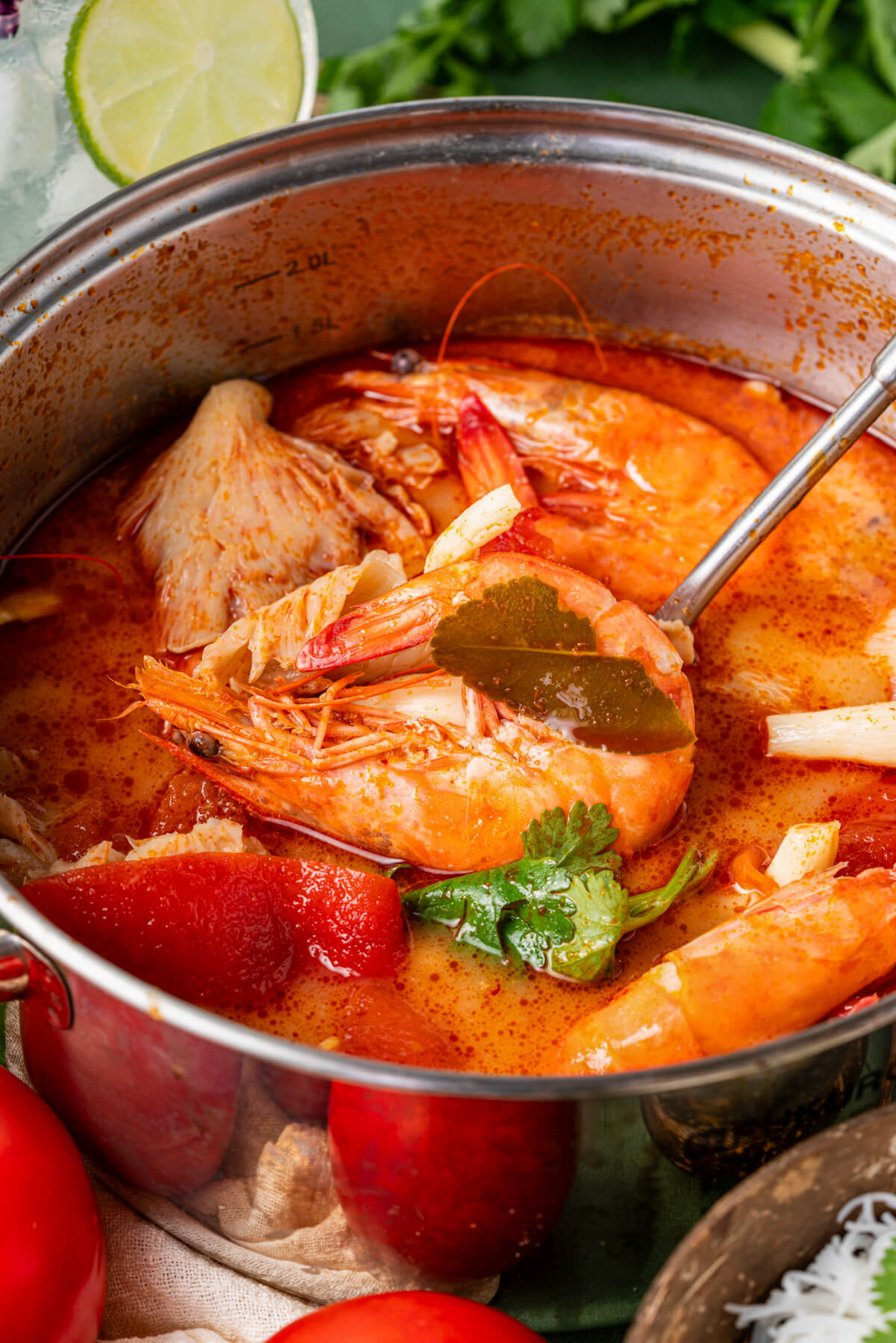 A ladle holding a giant prawn over a pot of vibrant red Tom Yum Soup.