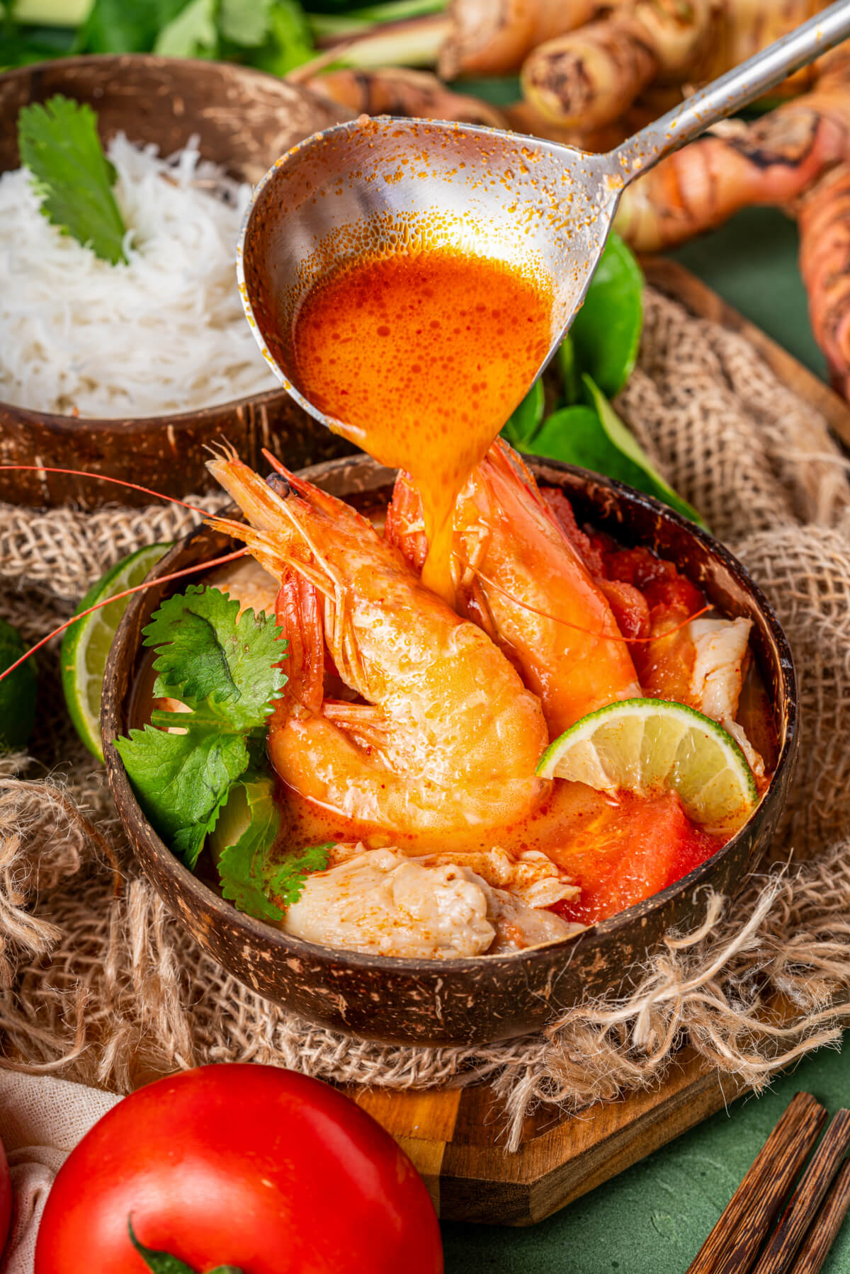 Vibrant red Tom Yum soup broth being poured over two cooked prawns in a bowl of soup garnished with fresh herbs and a lime wedge.